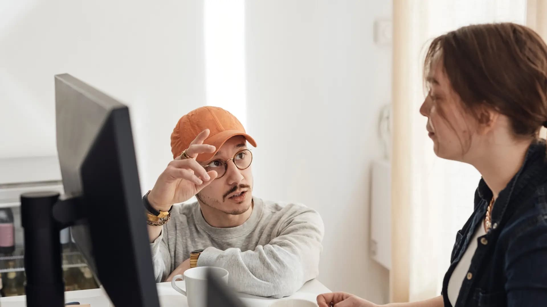 A woman sits in front of a monitor while a colleague kneels beside her chatting.