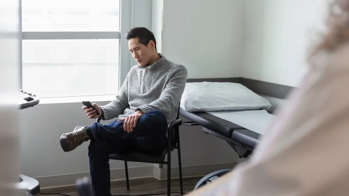 Man sitting in a doctor's examination room sits in a chair looking at his mobile device.